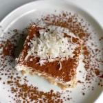 Square slice of Coconut Rum Coquito Tiramisu with creamy layers, dusted with cinnamon and topped with a generous mound of shredded coconut on a white plate, surrounded by cinnamon sprinkles, creating a festive and appealing presentation.