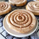 Extra-large Crumbl Churro Cookie with golden-brown edge and thick cinnamon frosting spiral, sprinkled with cinnamon-sugar, served on a white plate.