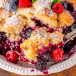 Close-up of Easy Berry Cobbler featuring golden, crumbly vanilla cake topping dusted with powdered sugar, vibrant mixed berries like raspberries, blueberries, and blackberries, all served in a white dish, highlighting its bright colors and inviting textures.