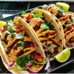 Three Garlic Butter Honey BBQ Chicken Tacos with golden-brown chicken, vibrant cilantro, crisp lettuce, red onion, creamy sauce drizzles, and lime wedges, arranged on a glossy black platter.