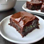 A rich, glossy Gluten Free Zucchini Brownie topped with thick chocolate frosting, showing fudgy texture and green zucchini flecks on a white plate