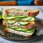 High-Protein Cucumber Sandwich with creamy dill spread, fresh cucumber ribbons, and whole wheat bread stacked on a black plate, garnished with fresh sprigs for a vibrant, appetizing presentation.