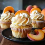 Moist Honey Peach Cream Cheese Cupcakes topped with creamy frosting, a drizzle of honey, and fresh peach slices on a black plate, with peach wedges for a vibrant, summery presentation.