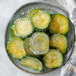 Vibrant green slices of frozen zucchini, glistening with icy frost and neatly arranged on a gray plate, showcasing their fresh summer color and smooth texture for how to freeze zucchini.