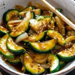 Glossy Japanese Zucchini wedges with onions, teriyaki glaze, and sesame seeds in a white bowl, showcasing green and golden hues and an appetizing, saucy presentation.