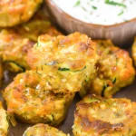 Crispy golden Keto Zucchini Tots with flecks of green zucchini and gooey melted cheese, arranged on a plate with a creamy dipping sauce in the background.