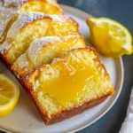 Moist lemon curd loaf cake with glossy lemon curd swirls, golden crumb, and a dusting of powdered sugar, beautifully sliced on a white plate with fresh lemon halves