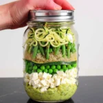 Bright, colorful layers of zucchini noodles, quinoa, asparagus, green peas, feta, and creamy dressing in a mason jar for Mason Jar Zucchini Noodle Salad with Coconut-Lime-Avocado Dressing.