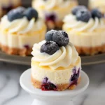 Mini Lemon Blueberry Cheesecakes topped with whipped cream and fresh blueberries, on golden graham crust, displayed on a white pedestal with a creamy, vibrant, and inviting appearance.
