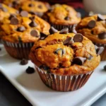 Moist Pumpkin Chocolate Chip Muffins with golden orange tops and rich chocolate chips, baked in paper liners on a white plate, showcasing their fluffy texture and inviting appearance.
