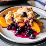 Golden, crisp-topped Peach Blueberry Cobbler with juicy peaches and blueberries bubbling beneath, served on a white plate with vibrant colors and textures, highlighting the inviting, fresh-baked presentation.