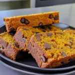 Moist pumpkin bread slices with rich orange color, studded with chocolate chips, arranged on a black plate, showcasing tender crumb and glossy finish