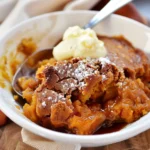 Golden pumpkin cobbler with a gooey caramel sauce, topped with powdered sugar and a scoop of vanilla ice cream in a white bowl.