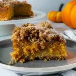 Golden, crumb-topped slice of Pumpkin Coffee Cake with moist orange interior, served on a gray plate beside a full cake and decorative pumpkins.