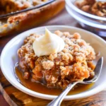 Golden Pumpkin Crisp with crumbly oat topping and creamy whipped topping, showing luscious orange filling, crispy clusters, and sticky syrup on a white bowl—an inviting autumn dessert.