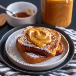 Golden Pumpkin Eggnog French Toast slices stacked on a plate, topped with creamy pumpkin butter and a dusting of powdered sugar, presented with a jar of pumpkin butter and striped napkin for a cozy, inviting breakfast scene.
