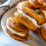 Golden pumpkin whoopie pies filled with creamy maple cream cheese, stacked on a bright white plate, showcasing their fluffy texture and inviting orange color.