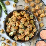 Golden, crispy Southern Fried Okra pieces with vibrant green centers in a rustic pan, surrounded by fresh whole okra and served with creamy dipping sauce.