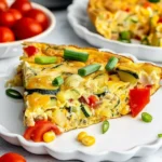 Golden crustless zucchini pie slice with vibrant red peppers, green zucchini, yellow corn, and melted cheddar, topped with fresh scallions on a white plate