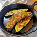 Glossy, caramelized Thomas Keller's Roasted Zucchini with golden brown glaze, sesame seeds, and vibrant sliced scallions in a black cast iron skillet