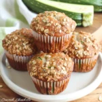 Four golden brown Zucchini Oatmeal Muffins topped with oats, in white liners, stacked on a white plate, with visible green zucchini flecks and a rustic, hearty texture.