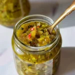 Colorful Zucchini Relish in a glass jar, featuring vibrant green zucchini, red pepper flecks, and a glossy, juicy texture, shown with an ornate golden spoon.
