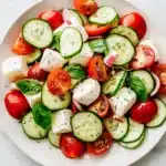 Colorful Fresh Cucumber Caprese Salad with mozzarella, tomatoes, cucumbers, onions, and basil on a white plate.