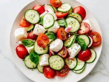 Colorful Fresh Cucumber Caprese Salad with mozzarella, tomatoes, cucumbers, onions, and basil on a white plate.