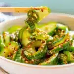 Bright, spiralized cucumber salad with sesame seeds and chili oil in a white bowl, being scooped up with chopsticks.