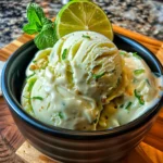 Vibrant green avocado ice cream in a black bowl, topped with creamy sauce, fresh mint, and a bright lime slice, with a smooth, inviting texture on a wooden board.