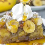 Golden caramel-glazed Banana Upside Down Cake slice with luscious banana rounds, glossy sauce, and a swirl of whipped cream on a white plate, dusted lightly with powdered sugar