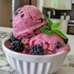 Glossy scoops of blackberry frozen yogurt, studded with whole blackberries and topped with fresh green mint, in a white bowl on a marble countertop.