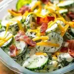 Creamy cucumber salad with bacon and cheddar cheese in a clear bowl, garnished with fresh dill and vibrant cheese shreds.
