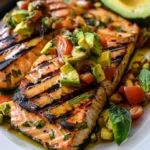 Vibrant grilled salmon fillets with dark char lines topped with colorful avocado bruschetta and cherry tomatoes, garnished with fresh basil, on a white platter.