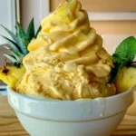 Creamy, golden-yellow Homemade Dole Whip swirled high in a white bowl, garnished with fresh pineapple wedges and vibrant green mint, radiating tropical freshness and soft, velvety texture.