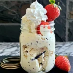 Creamy Mason Jar Vanilla Bean Ice Cream with visible vanilla specks, topped with whipped cream and fresh strawberries, beautifully presented in a clear glass mason jar on marble with extra berries on the side.