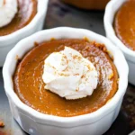 Rich orange Mini Keto Crustless Pumpkin Pie in white ramekin, topped with fluffy whipped cream and a dusting of cinnamon, glistening with a glossy, creamy finish.