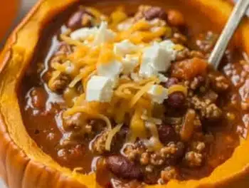 Vibrant orange Pumpkin Chili served in a pumpkin bowl, topped with melty cheddar and creamy cheese, highlighting rich beans and hearty turkey in a savory, colorful presentation with a silver spoon.