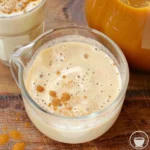 Smooth and creamy Pumpkin Spice Coffee Creamer in a glass pitcher, topped with a sprinkle of pumpkin spice, next to a jar of golden pumpkin mixture on a rustic wooden table.