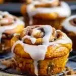 Golden pumpkin cinnamon roll muffins with glossy cream cheese glaze dripped over deep orange swirls, served on a wire rack, showcasing vibrant color and inviting texture.
