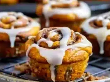 Golden pumpkin cinnamon roll muffins with glossy cream cheese glaze dripped over deep orange swirls, served on a wire rack, showcasing vibrant color and inviting texture.
