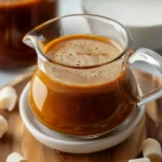 Glossy golden-brown Vanilla Pumpkin Marshmallow Coffee Syrup in a clear glass pitcher, surrounded by white marshmallows, on a wooden tray with a smooth, creamy texture and inviting autumn warmth.