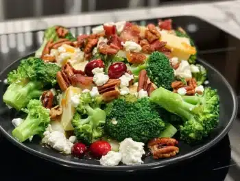 A vibrant broccoli salad on a black plate, topped with toasted pecans, bacon, dried cranberries, feta, and cheddar.