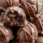 Chocolate-covered no-bake brownie batter protein bites with creamy chocolate centers and a glossy drizzle, stacked on a plate.