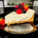 A slice of creamy no-bake vanilla cheesecake on a black plate garnished with fresh strawberries, blackberries, and raspberries.