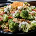 Closeup of vibrant Easy Broccoli Salad piled high on a black plate, topped with golden raisins, crisp bacon, toasted almonds, shredded cheddar, and creamy dressing.