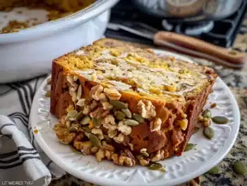 Close-up of a slice of homemade pumpkin bread topped with chopped walnuts and pumpkin seeds on a white plate