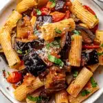 Close-up of vibrant roasted eggplant pasta with cherry tomatoes and fresh basil on a white plate